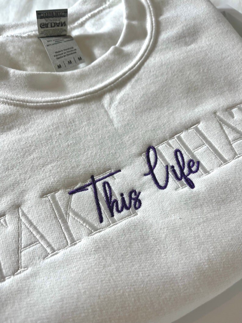 K&ouml;nnte beinhalten: Wei&szlig;es Sweatshirt mit dem gestickten Text "TAKE THIS LIFE" in Lila. Das Sweatshirt hat einen Rundhalsausschnitt und ein Etikett am inneren Kragen. Der Stoff wirkt weich und bequem.