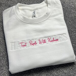 May include: A white sweatshirt featuring the embroidered words "HAPPY PEOPLE With Kindness" in pink. The crew neck sweatshirt has long sleeves. The size tag reads "M".