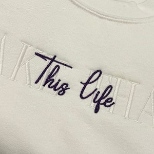 K&ouml;nnte beinhalten: Ein wei&szlig;es Sweatshirt mit dem gestickten Schriftzug "Take this life that". Die W&ouml;rter "Take" und "life" sind in dunkellila Schrift, w&auml;hrend "this" und "that" in wei&szlig;er Blockschrift gehalten sind.