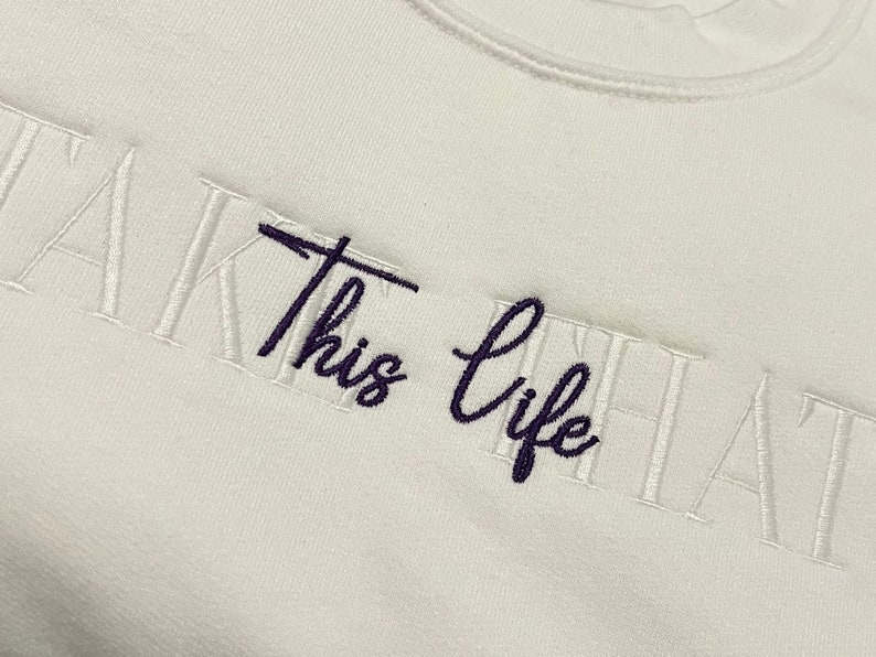 K&ouml;nnte beinhalten: Ein wei&szlig;es Sweatshirt mit dem gestickten Schriftzug "Take this life that". Die W&ouml;rter "Take" und "life" sind in dunkellila Schrift, w&auml;hrend "this" und "that" in wei&szlig;er Blockschrift gehalten sind.