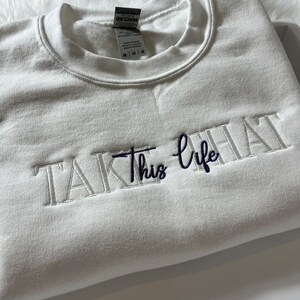 K&ouml;nnte beinhalten: Ein wei&szlig;es Sweatshirt mit dem gestickten Schriftzug "TAKE this life" in wei&szlig;er und marineblauer Farbe. Das Sweatshirt ist gefaltet und scheint aus einem weichen Material zu sein.