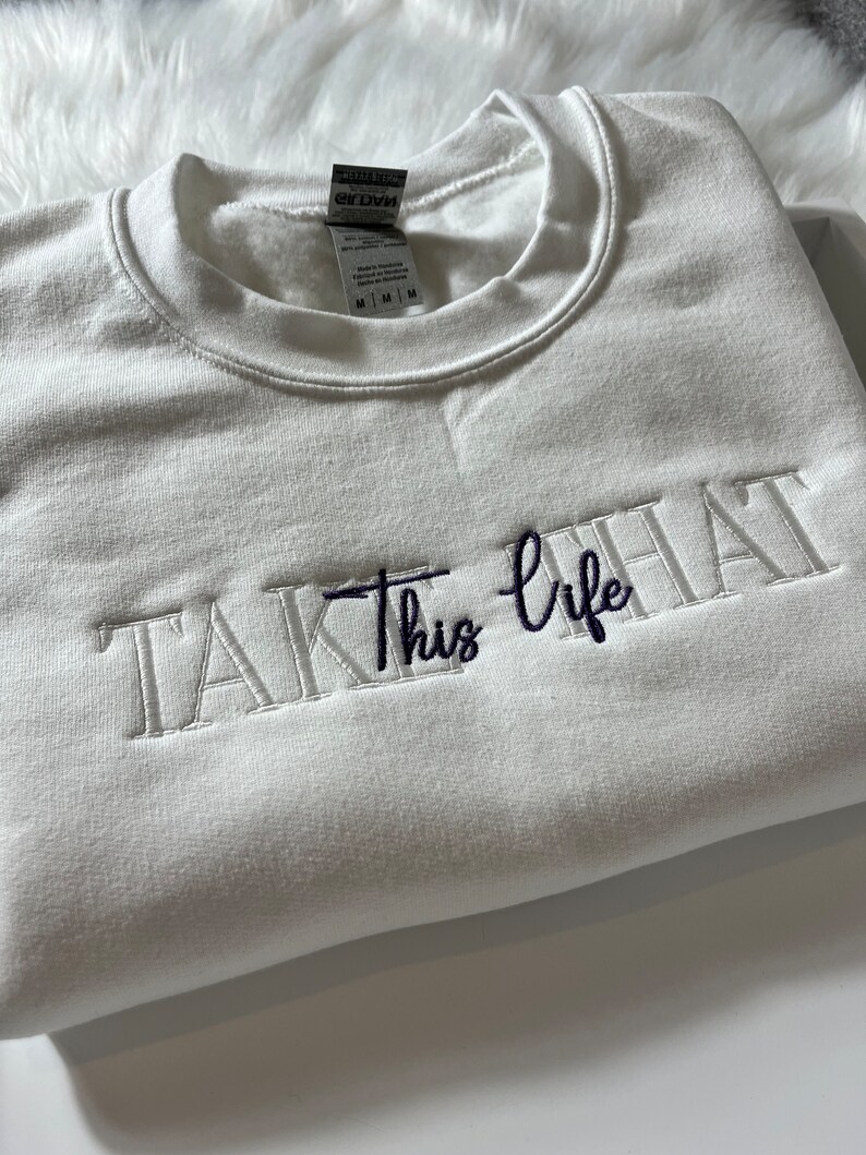 K&ouml;nnte beinhalten: Ein wei&szlig;es Sweatshirt mit dem gestickten Schriftzug "TAKE this life" in wei&szlig;er und marineblauer Farbe. Das Sweatshirt ist gefaltet und scheint aus einem weichen Material zu sein.