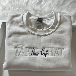 K&ouml;nnte beinhalten: Ein gefalteter wei&szlig;er Sweatshirt mit dem gestickten Schriftzug "TAKE This life" in einer Mischung aus Serifen- und Schreibschriften. Der Sweatshirt ist aus einem weichen Material und ist sauber gefaltet. Das Etikett ist sichtbar.