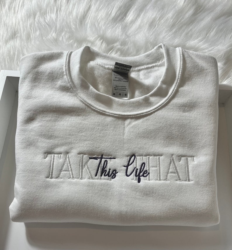 K&ouml;nnte beinhalten: Ein gefalteter wei&szlig;er Sweatshirt mit dem gestickten Schriftzug "TAKE This life" in einer Mischung aus Serifen- und Schreibschriften. Der Sweatshirt ist aus einem weichen Material und ist sauber gefaltet. Das Etikett ist sichtbar.