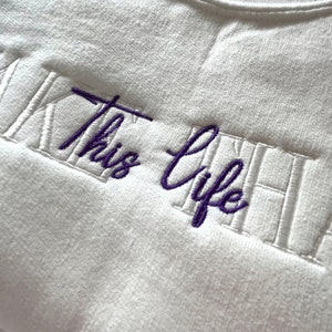 K&ouml;nnte beinhalten: Ein wei&szlig;es Sweatshirt mit Rundhalsausschnitt und dem gestickten Text "This Life" in Lila. Der Text ist zentriert und in einer Schreibschrift gestickt, mit einem rechteckigen Rahmen. Das Sweatshirt hat einen gerippten Ausschnitt.