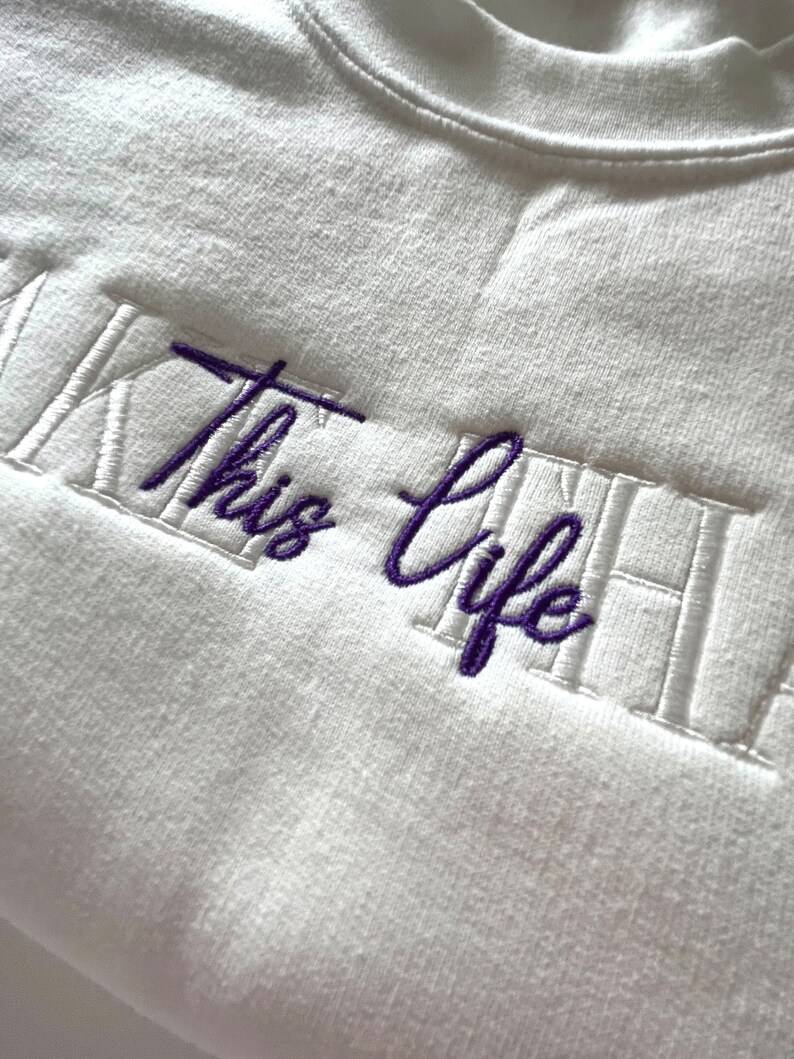 K&ouml;nnte beinhalten: Ein wei&szlig;es Sweatshirt mit Rundhalsausschnitt und dem gestickten Text "This Life" in Lila. Der Text ist zentriert und in einer Schreibschrift gestickt, mit einem rechteckigen Rahmen. Das Sweatshirt hat einen gerippten Ausschnitt.