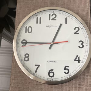 May include: A round, silver-framed wall clock with a white face. The clock features black numerals, black and red hands, and the word "skytime" above the center. The word "QUARTZ" is printed below the center.
