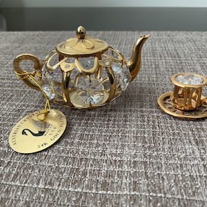 May include: A miniature gold-plated teapot and teacup set with clear crystal accents. The teapot has a handle, spout, and lid. The teacup sits on a matching saucer. A gold tag reads "Austrian Crystal" and "24K Gold Plated."