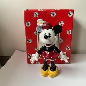 May include: A Minnie Mouse figurine stands in front of a red box. The figurine is white, black, and red, with yellow shoes. The box is red with white circles and Mickey Mouse graphics.
