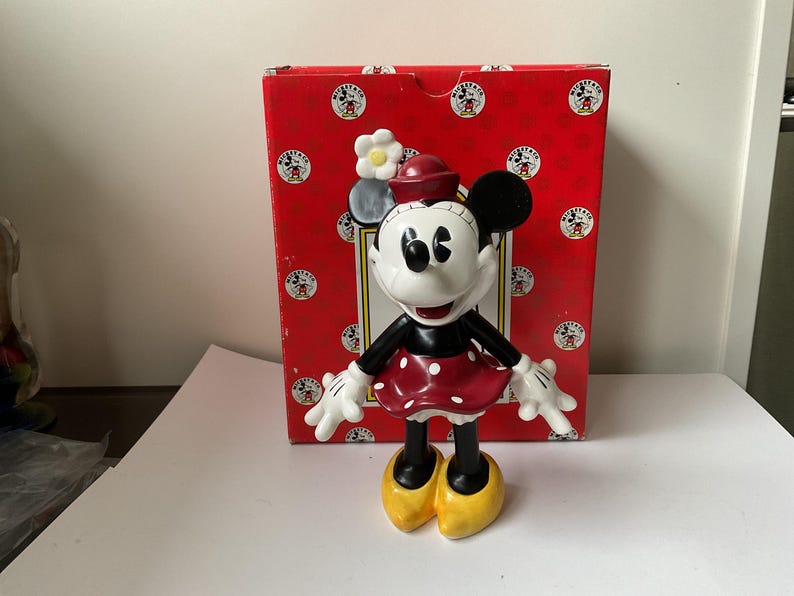 May include: A Minnie Mouse figurine stands in front of a red box. The figurine is white, black, and red, with yellow shoes. The box is red with white circles and Mickey Mouse graphics.