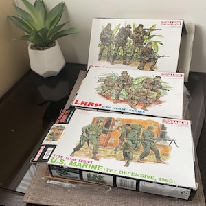 May include: Three model kit boxes with illustrations of soldiers in combat. The boxes are labelled with text including "LRRP", "U.S. MARINE", and "TET OFFENSIVE, 1968". The kits are from the "Dragon" brand and are 1/35 scale.