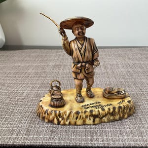 May include: A vintage figurine of a fisherman holding a fishing rod, wearing a hat and traditional clothing. The figurine is on a base with a teapot, fish, and text that reads "CANADIAN NATIONAL EXHIBITION 1965".