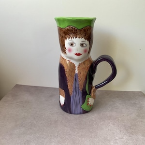 May include: A ceramic jug shaped like a person with brown hair, wearing a purple coat with a tan fur-like collar and a green bag. The jug features a green rim, a purple handle, and a painted face with red lips and rosy cheeks.