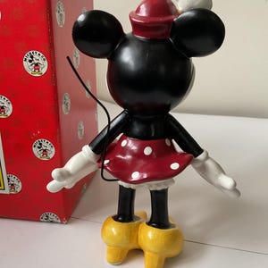May include: A black Minnie Mouse figurine with a red and white polka dot skirt, white gloves, and yellow shoes. The figurine has a red hat and a white chef's hat. A red box with the Mickey & Co. logo is in the background.
