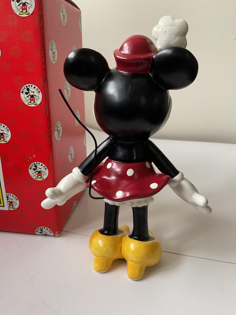 May include: A black Minnie Mouse figurine with a red and white polka dot skirt, white gloves, and yellow shoes. The figurine has a red hat and a white chef's hat. A red box with the Mickey & Co. logo is in the background.