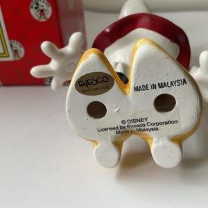 May include: A ceramic Mickey Mouse figurine with a yellow and white base, red shorts, and white gloves. The figurine has the text "MADE IN MALAYSIA" and "&copy; DISNEY" printed on the bottom. A red box with Mickey Mouse graphics is in the background.