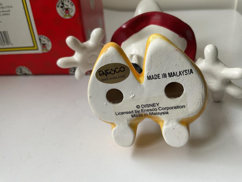 May include: A ceramic Mickey Mouse figurine with a yellow and white base, red shorts, and white gloves. The figurine has the text "MADE IN MALAYSIA" and "&copy; DISNEY" printed on the bottom. A red box with Mickey Mouse graphics is in the background.