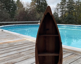 May include: A vertical wooden shelf shaped like a boat hull with a dark brown stained finish. The structure features a pointed top and two horizontal shelves inside, creating three distinct storage sections within the curved wooden frame.