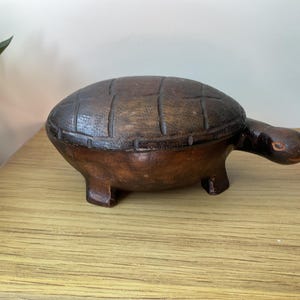 May include: A dark brown wooden turtle-shaped box with a removable lid. The shell features a carved grid pattern, and the head has small, painted orange eyes. The box stands on four short, sturdy legs and has a smooth, polished finish throughout.