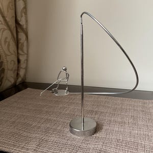 May include: A silver metal sculpture of a person holding a fishing rod, with a curved metal rod forming an arc above the person. The sculpture is on a brown textured surface.