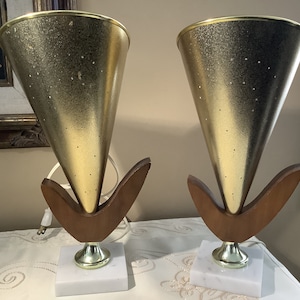 Könnte beinhalten: Zwei Tischlampen im Stil der modernen Mitte des Jahrhunderts mit goldenen Kegel-Lampenschirmen und Holzsockeln. Die Lampenschirme haben eine gesprenkelte goldene Oberfläche und sind mit kleinen Löchern verziert. Die Sockel sind aus dunklem Holz gefertigt und haben ein einzigartiges, geschwungenes Design. Die Lampen stehen auf weißen Marmorsockeln.