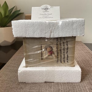 Könnte beinhalten: Eine hölzerne Erinnerungsbox mit einem kleinen gerahmten Bild einer Figur, die ein Herz hält. Die Box ist in klarem Kunststoff verpackt und von weißem Schaumstoff umgeben. Eine kleine Karte obenauf lautet "From the Heart, Love, heartfelt and true."