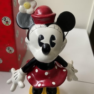 May include: A Minnie Mouse figurine with a red dress with white polka dots, yellow shoes, and a red hat. The figurine has a white flower on her head and black ears. A red box with Mickey Mouse graphics is in the background.