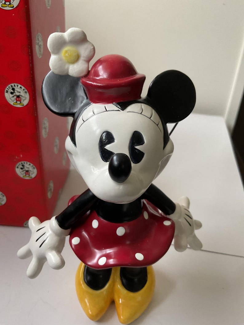 May include: A Minnie Mouse figurine with a red dress with white polka dots, yellow shoes, and a red hat. The figurine has a white flower on her head and black ears. A red box with Mickey Mouse graphics is in the background.