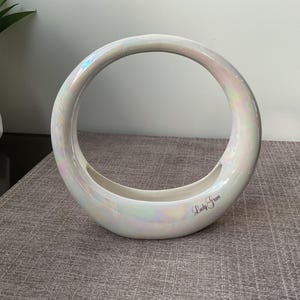 Pode incluir: Um vaso em forma de anel de cerâmica branca perolada com uma base larga e uma abertura circular. O vaso tem um acabamento brilhante e um brilho iridescente. O texto "Lady Grace" está escrito na lateral. O vaso está sobre uma superfície cinza texturizada.