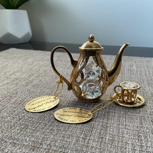 May include: A gold-plated teapot and teacup set with clear crystal accents. The teapot has a decorative openwork design, and the teacup sits on a matching saucer. The set includes two gold tags with text. A decorative item.