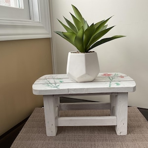 May include: A small, white, wooden plant stand with a floral design on the top surface. A white geometric planter holds a green plant. The stand is painted white with a distressed finish.