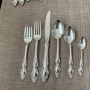 Pode incluir: Um conjunto de talheres prateados, incluindo garfos, uma faca e colheres, dispostos sobre uma superfície castanha. Os talheres têm detalhes ornamentados, semelhantes a flores, nas pegas. O conjunto inclui um garfo de jantar, um garfo de salada, uma faca, uma colher de sopa, uma colher de chá e uma colher menor.