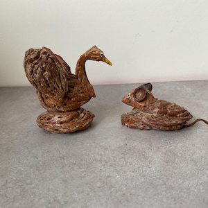 Könnte beinhalten: Zwei handgefertigte Figuren aus strukturierten braunen Walnussschalen. Eine Figur stellt einen Schwan mit langem Hals und gelbem Schnabel dar, während die andere eine Maus mit abgerundeten Ohren und einem dünnen, geschwungenen Schwanz zeigt.