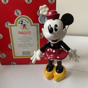 May include: A Minnie Mouse figurine with a red polka-dot dress, black arms and legs, and yellow shoes. The figurine has a red hat and white gloves. The box behind the figurine is red with Mickey Mouse logos and the text "Mickey & Co."