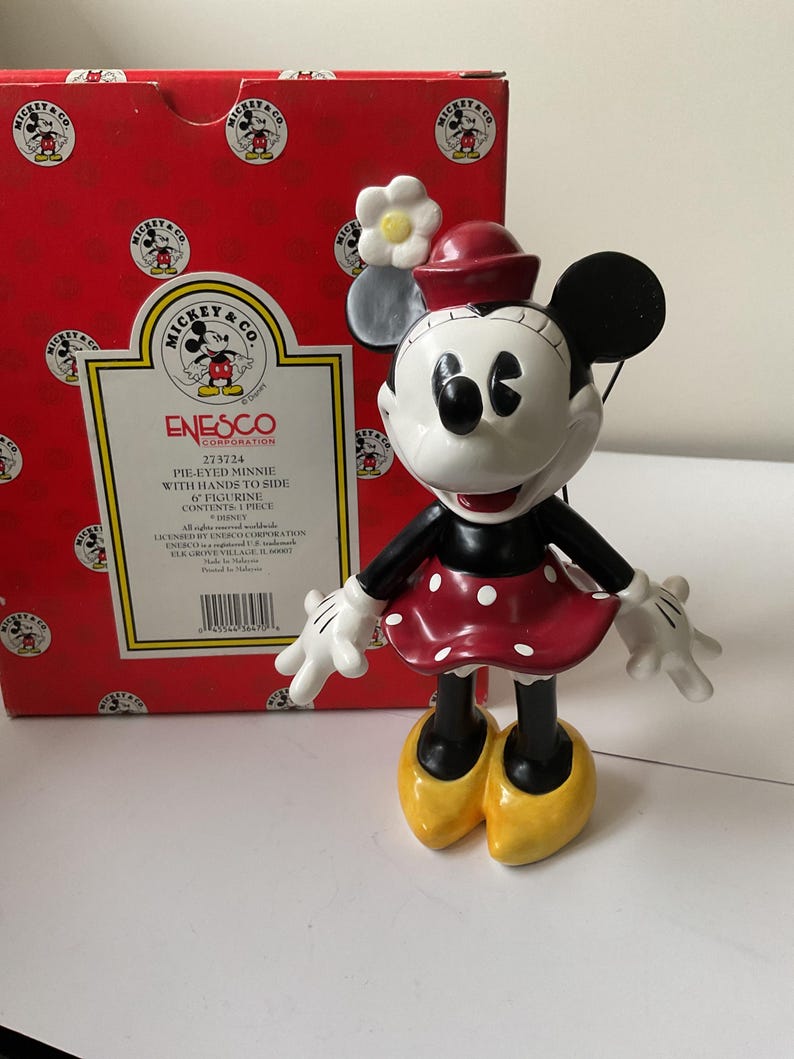 May include: A Minnie Mouse figurine with a red polka-dot dress, black arms and legs, and yellow shoes. The figurine has a red hat and white gloves. The box behind the figurine is red with Mickey Mouse logos and the text "Mickey & Co."