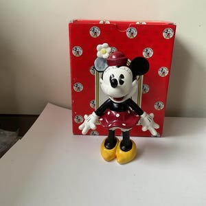May include: A Minnie Mouse figurine with a red polka-dot dress, black gloves, and yellow shoes. The figurine has a red hat with a white flower and is standing in front of a red box with Mickey Mouse logos.