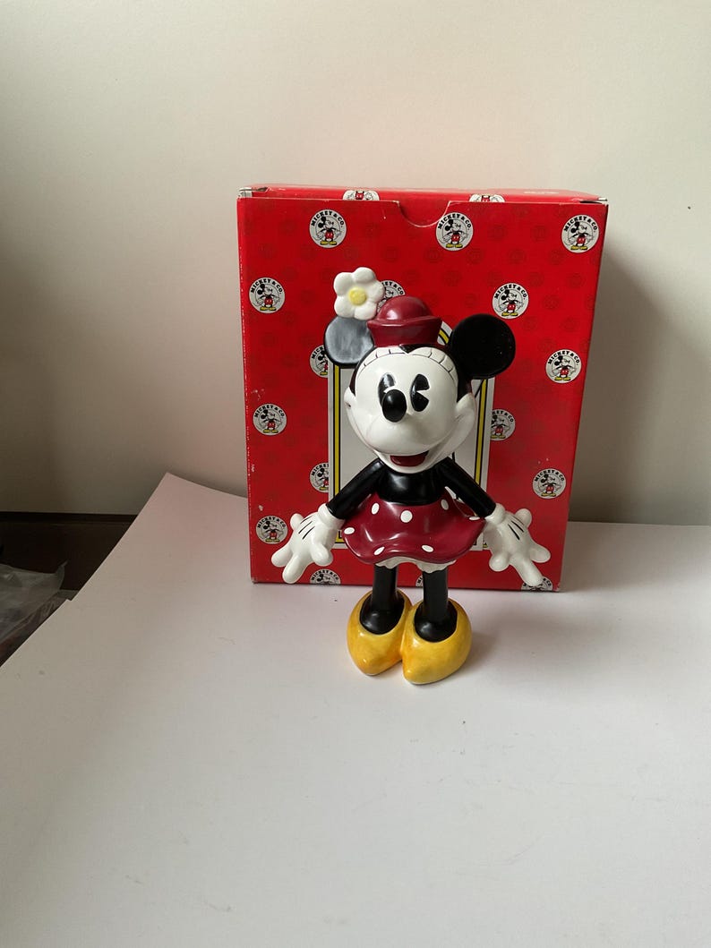 May include: A Minnie Mouse figurine with a red polka-dot dress, black gloves, and yellow shoes. The figurine has a red hat with a white flower and is standing in front of a red box with Mickey Mouse logos.