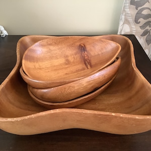 Pode incluir: Um conjunto de tigelas de madeira para servir. O conjunto inclui uma tigela quadrada grande com bordas arredondadas e três tigelas ovais menores aninhadas dentro. A madeira tem um tom quente e natural.