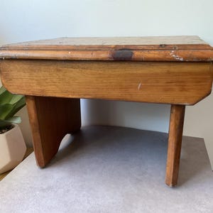 May include: A small, rectangular wooden stool with a natural light brown finish. The stool features a flat top surface with visible wood grain and wear, supported by one solid panel leg on the left and one cylindrical leg on the right side.
