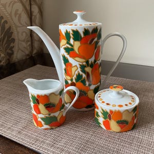 May include: A white ceramic teapot, creamer and sugar bowl with a floral design. The teapot has a handle and spout. The creamer and sugar bowl have lids. The floral design features orange, green and yellow flowers on a white background.