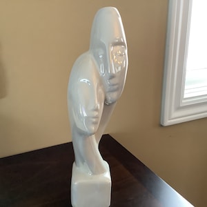 May include: White ceramic sculpture of two abstract faces, one face is partially obscured by the other. The sculpture is on a square base.