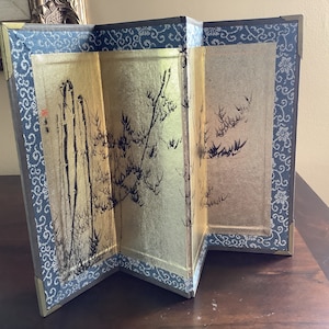Peut inclure: Petit paravent pliable avec des panneaux dorés ornés d'illustrations à l'encre noire de bambous et de plantes. Bordure bleu foncé avec motif floral blanc et coins en laiton.