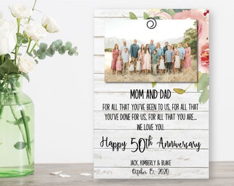 May include: A white wood sign with a photo of a family in front of a mountain range. The sign has the text "Mom and Dad" and "Happy 50th Anniversary" with a floral design. The text "Jack, Kimberly & Blake" and "October 15, 2020" is also included.