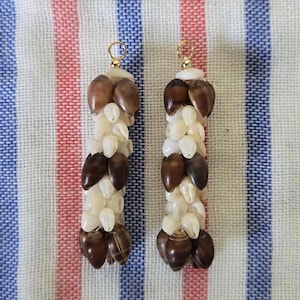 May include: Two handmade earrings composed of small, white and brown seashells. The shells are strung together in a vertical pattern, with gold-colored loops at the top. The earrings are displayed on a striped fabric background.