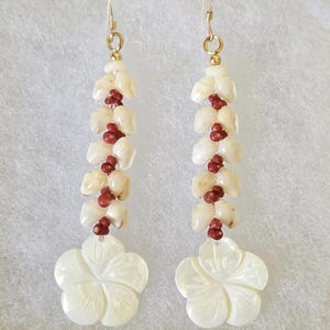 May include: A pair of white shell earrings with red beads and a carved hibiscus flower design. The earrings are displayed in a white box with a gold trim.