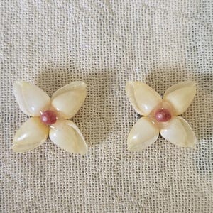 May include: Two flower-shaped earrings. Each earring has four off-white petals surrounding a small, pink center. The earrings are set against a textured, off-white fabric background. The earrings are likely for pierced ears.