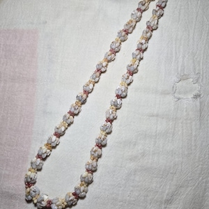 May include: A delicate necklace composed of small, white shell-like beads, interspersed with tiny pink and yellow beads. The necklace is laid out on a white fabric surface, showcasing its intricate design and natural color palette.