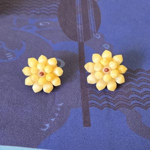 May include: A pair of yellow flower-shaped earrings. Each earring features a central cluster of small, round beads with a reddish-brown center, surrounded by petal-shaped elements. The earrings are set against a blue patterned background.