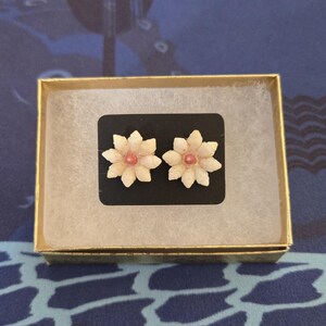May include: A pair of flower-shaped stud earrings with a light beige color and a pink center, presented in a gold box with a black backing. The earrings are delicate and detailed, resembling small blossoms.