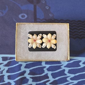 May include: A pair of flower-shaped earrings made from seashells, presented in a small gold-trimmed box. Each earring features a central red bead surrounded by cream-colored petals and shell accents, set on a black base.
